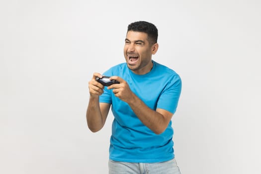 Portrait of excited extremely happy unshaven man gamer wearing blue T- shirt standing playing video games, holding joystick in hands. Indoor studio shot isolated on gray background.