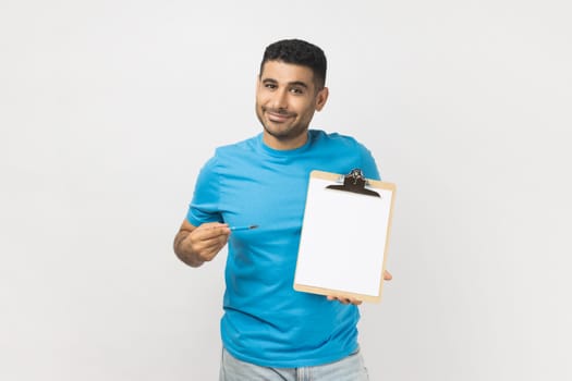 Portrait of smiling happy delighted unshaven man wearing blue T- shirt standing pointing at clipboard with empty paper, copy space. Indoor studio shot isolated on gray background.