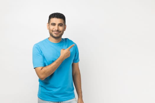 Portrait of smiling cheerful attractive unshaven man wearing blue T- shirt standing pointing copy space for advertisement or promotional text. Indoor studio shot isolated on gray background.