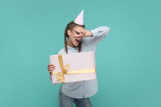 Portrait of joyful beautiful teenager girl wearing striped shirt and party cone, holding present box, showing v sign, celebrating her birthday. Indoor studio shot isolated on green background.