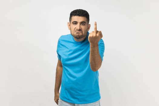 Sign of protest. Portrait of vulgar unshaven man wearing blue T- shirt shows middle finger impolite gesture disrespects someone looks rude at camera. Indoor studio shot isolated on gray background.