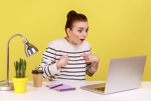 Surprised woman with shocked expression looking at laptop screen with open mouth, talking on video call, online conference from home office. Indoor studio studio shot isolated on yellow background.