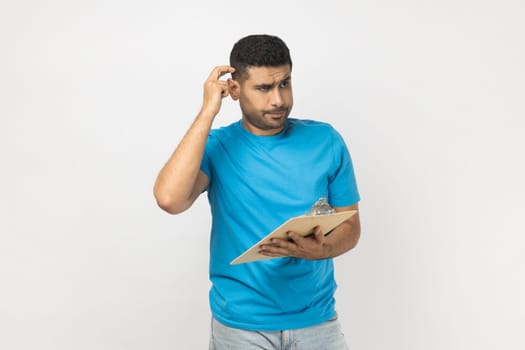 Portrait of pensive puzzled unshaven man wearing blue T- shirt standing with clipboard, writing something on paper, thinking about plan. Indoor studio shot isolated on gray background.