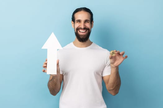 Portrait of smiling dark haired man with beard wearing white T-shirt showing growth increase of bitcoin, expressing positive emotions. Indoor studio shot isolated on blue background.