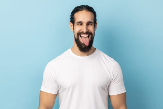 Portrait of comic positive man with beard wearing white T-shirt looking cross-eyed, having fun with silly face expression, showing tongue out. Indoor studio shot isolated on blue background.