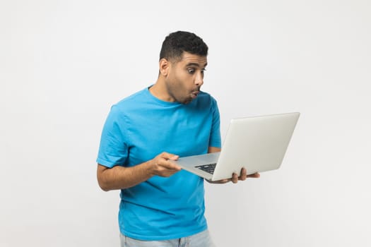 Portrait of surprised astonished unshaven man wearing blue T- shirt standing holding laptop in hands, working online, looking at screen with big eyes. Indoor studio shot isolated on gray background.