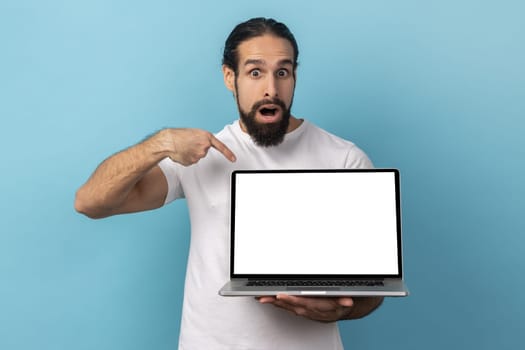 Portrait of man with beard wearing white T-shirt standing holding laptop with blank screen and looking at camera with open mouth, internet advertising. Indoor studio shot isolated on blue background.