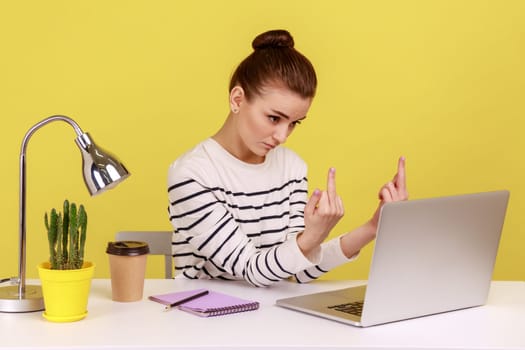 Rude woman office manager showing middle finger to laptop screen, aggressively communicating on video call, expressing disrespect and hate. Indoor studio studio shot isolated on yellow background.