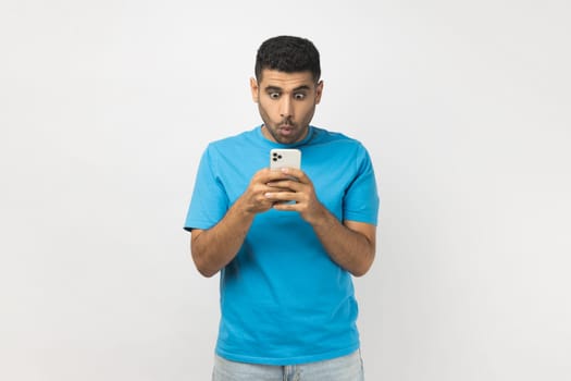 Portrait of shocked amazed unshaven man wearing blue T- shirt standing using mobile phone, checking social networks, reading news. Indoor studio shot isolated on gray background.