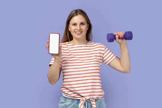 Portrait of delighted good looking woman wearing striped T-shirt holding dumbbell in hand, showing smart phone with empty display for advertisement. Indoor studio shot isolated on purple background.