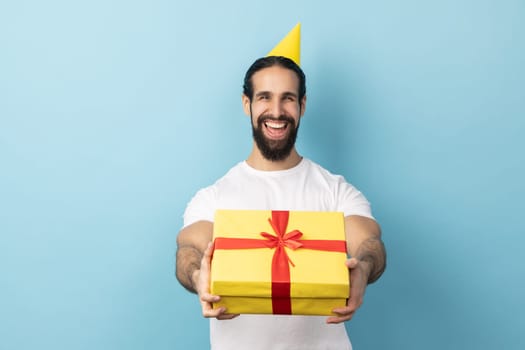 Portrait of man with beard wearing white T-shirt and party cone giving present and congratulating on birthday, offering gift box for holiday. Indoor studio shot isolated on blue background.