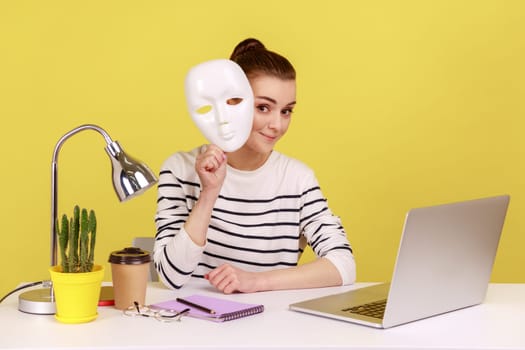 Woman with dark hair removing white mask from face showing his smiling expression while sitting at workplace with laptop, good mood. Indoor studio studio shot isolated on yellow background.