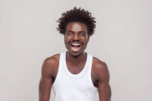 Emotional gorgeous man with Afro hairstyle grinning broadly, enjoying good day, having fun, laughing at something funny, wearing white T-shirt. Indoor studio shot isolated on gray background.