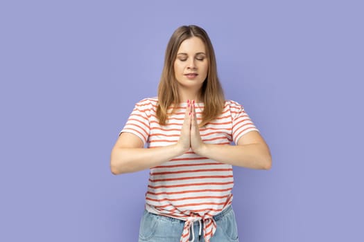 Portrait of blond woman wearing striped T-shirt standing in yoga pose with closed eyes and try to relaxing, keeps palms together. Indoor studio shot isolated on purple background.