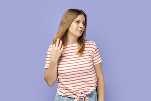 Portrait of charming inspired blond woman wearing striped T-shirt looking away with pensive expression, dreaming pleasant thoughts. Indoor studio shot isolated on purple background.