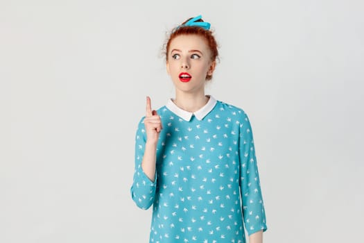 Portrait of excited clever smart red haired woman with bun hairstyle, standing raised index finger up, having idea, looking away, wearing blue dress. Indoor studio shot isolated on gray background.