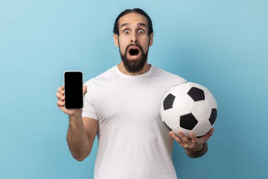 Portrait of amazed man with beard wearing white T-shirt holding soccer ball and smartphone empty black display, ticket booking for championship. Indoor studio shot isolated on blue background.