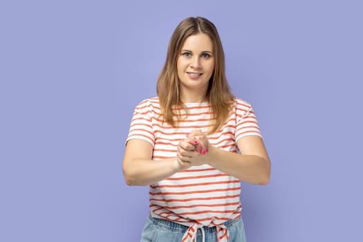 Portrait of winsome positive delighted woman wearing striped T-shirt standing with hands together, looking at camera with happy facial expression. Indoor studio shot isolated on purple background.