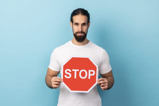 Portrait of serious man with beard wearing white T-shirt holding red Stop sign, looking at camera with negative aggressive expression, showing ban. Indoor studio shot isolated on blue background.