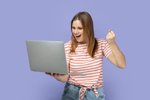 Portrait of extremely happy beautiful blond woman wearing striped T-shirt working on laptop, looking at screen and clenched fist, showing yes gesture. Indoor studio shot isolated on purple background.