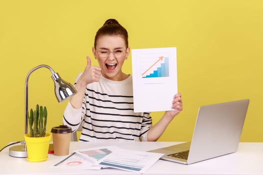 Delighted woman smiling at camera holding growth diagram, showing thumb up, satisfied with financial and economic growth of his business. Indoor studio studio shot isolated on yellow background.