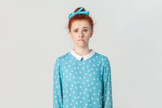 Portrait of scared frighten ginger woman with bun hairstyle, looking at camera and biting her lips, being nervous, wearing blue dress. Indoor studio shot isolated on gray background.