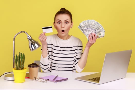 Electronic money. Shocked man sitting office workplace with laptop, holding dollars and credit card, looking at camera with open mouth. Indoor studio studio shot isolated on yellow background.