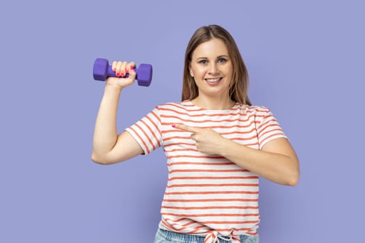 Portrait of satisfied delighted sporty blond woman wearing striped T-shirt holding dumbbell in hand, female pointing at her biceps and smiling. Indoor studio shot isolated on purple background.