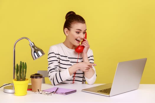 Woman secretary making calls on landline telephone working on laptop sitting at office, looking and pointing at notebook display. Indoor studio studio shot isolated on yellow background.