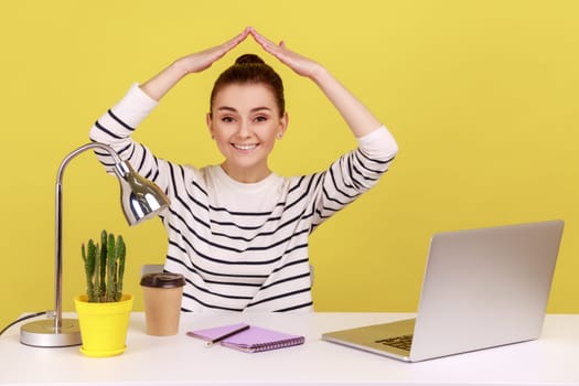 Protected life, safety. Cheerful positive woman doing house roof gesture over head, sitting at workplace with laptop, home office. Indoor studio studio shot isolated on yellow background.