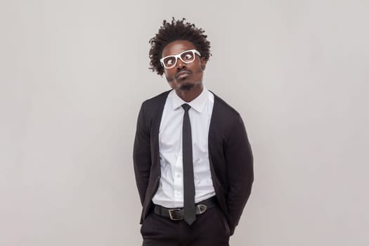 Portrait of funny comic man in glasses crosses eyes, pouts lips, makes grimace, foolishes after all day working, wearing white shirt and tuxedo. Indoor studio shot isolated on gray background.