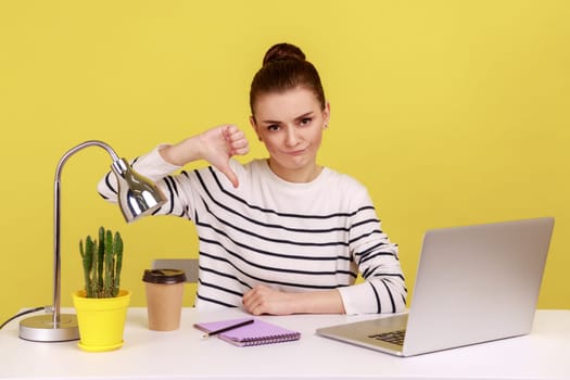 Displeased woman office worker showing thumbs down, dislike gesture, expressing disapproval, criticizing sitting at workplace with laptop. Indoor studio studio shot isolated on yellow background.