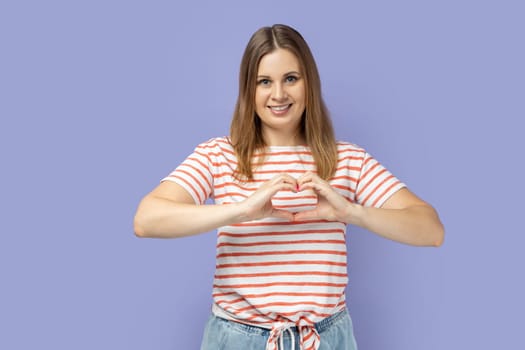Portrait of blond woman wearing striped T-shirt holding hands in shape of heart showing romantic gesture, love confession, valentines day celebration. Indoor studio shot isolated on purple background.