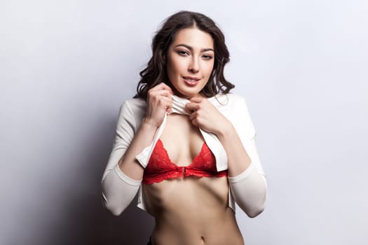 Portrait of smiling happy beautiful sexy woman with wavy hair taking off her shirt showing her bare belly and red underwear. Indoor shot isolated on gray background.