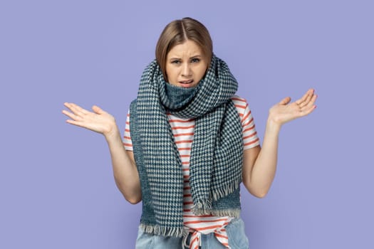 Portrait of confused helpless sick woman wearing striped T-shirt and wrapped in scarf standing with raised arms, catching cold, having flu symptoms. Indoor studio shot isolated on purple background.