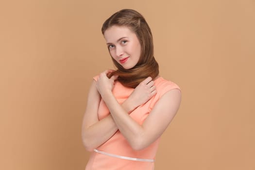 Portrait of cute charming attractive woman standing wrapped her neck with her long hair, looking at camera with flirting look, wearing elegant dress. Indoor studio shot isolated on brown background.
