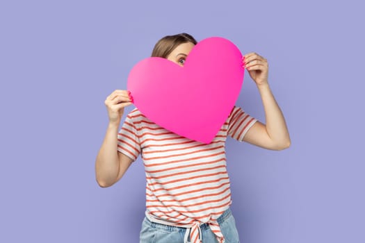 Portrait of shy flirting blond woman wearing striped T-shirt holding big pink heart and peeping out from love symbol, looking at camera. Indoor studio shot isolated on purple background.