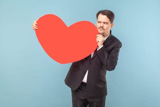 Portrait of confused cunning man with mustache standing with big red heart in hands, looking away, wearing black suit with red tie. Indoor studio shot isolated on light blue background.