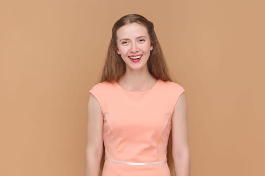 Portrait of joyful cheerful beautiful woman with long hair looking at camera with toothy smile, being in good mood, wearing elegant dress. Indoor studio shot isolated on brown background.