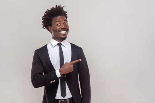 Man with Afro hairstyle has happy expression, smiles gently, points aside with fore finger, shows place to hang out, wearing white shirt and tuxedo. Indoor studio shot isolated on gray background.