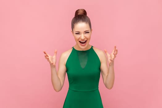 Portrait of crazy angry woman standing with raised arms, screaming with aggression, having problems, shouting, wearing green dress. Indoor studio shot isolated on pink background.