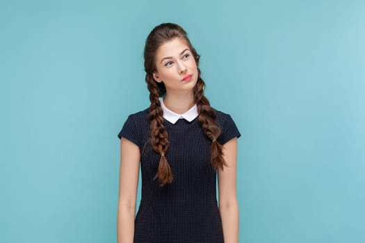 Portrait of pensive thoughtful woman with braids standing and looking away, making decision, thinks about solving problem, wearing black dress. woman Indoor studio shot isolated on blue background.