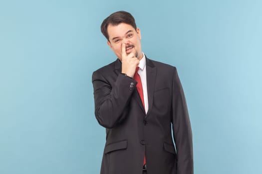 Portrait of funny childish man with mustache standing having comical facial expression, keeps finger in nose, wearing black suit with red tie. Indoor studio shot isolated on light blue background.