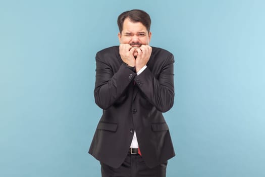 Portrait of nervous worried man with mustache standing biting his fingernails, having serious problems, scared, wearing black suit with red tie. Indoor studio shot isolated on light blue background.