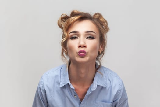 Air kiss for you. Portrait of blonde woman keeps lips folded, expressing love, blowing mwah, having romantic expression, wearing blue shirt. Indoor studio shot isolated on gray background.