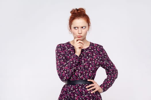 Portrait of redhead woman wearing dress holding chin and pondering idea, planning strategy, musing and solving difficult question in mind. Indoor studio shot isolated on gray background.