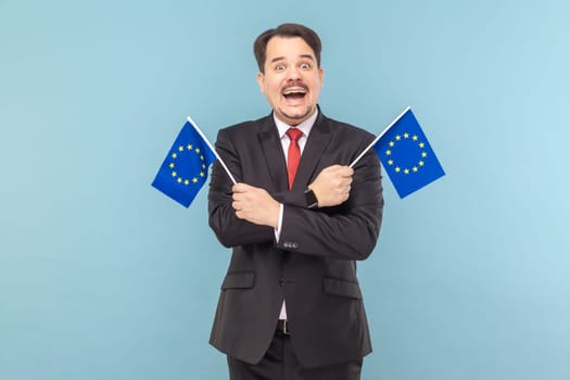 Man smiling broadly and holding flags of European Union, symbol of Europe, EU association and community, wearing black suit with red tie. Indoor studio shot isolated on light blue background.