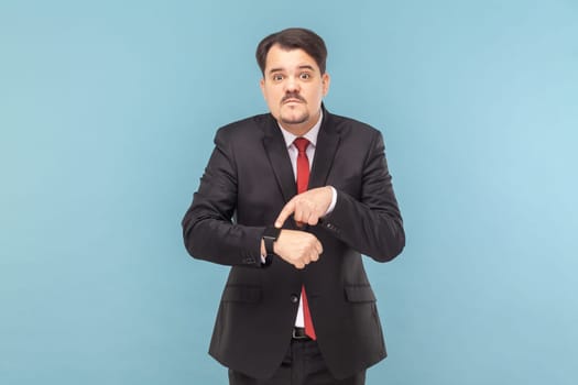 Portrait of serious man with mustache standing looking at camera, pointing at his smartwatch, deadline, wearing black suit with red tie. Indoor studio shot isolated on light blue background.