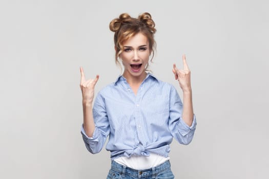 Portrait of blonde woman showing rock and roll gesture heavy metal sign enjoying favorite music on party has fun, exclaims from joy, wearing blue shirt. Indoor studio shot isolated on gray background.