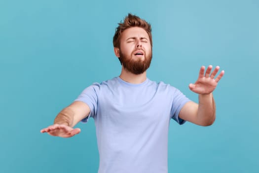 Loneless and blindness. Portrait of sick bearded man standing with closed eyes and try to touching something or find, posing with outstretching hands. Indoor studio shot isolated on blue background.